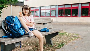 Zu sehen ist ein Mädchen, das alleine auf einer Bank auf einem verwaisten Schulhof sitzt.