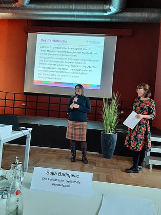 Vortrag, links Sabine Bahr mit Mikrofon, rechts Sejla Badnjevic. Im Hintergrund ist eine Leinwand mit Folie und Inhalt zum Paritätischen.