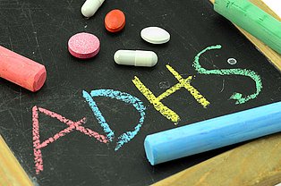 Schriftzug ADHS auf Tafel geschrieben, ein Kreidestück und ein paar Tabletten daneben liegend