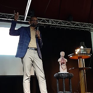 Zu sehen ist Dr. Etoung auf der Bühne mit Mikrofon in der Hand. Hinter ihm ist eine Leinwand. Auf der rechten Seite steht ein Thorax.