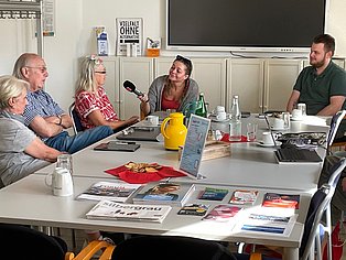 Foto von Menschen die an einem Tisch sitzen, links zwei hochaltrigere Frauen und ein Mann, vor Kopf eine jüngere Frau mit einem Mikro und re